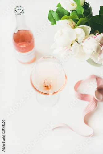 Rose wine in glass and bottle  pink decorative ribbon  peony flowers over white background  copy space. Summer celebration  wedding greeting card  invitation concept