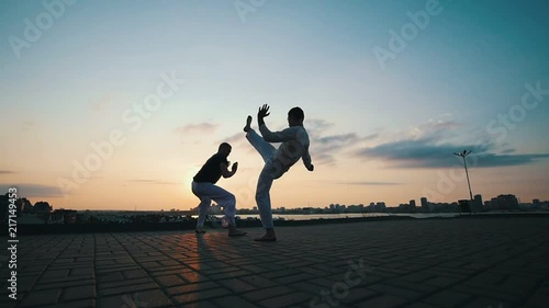 Men with a sporty physique dancing capoeira outdoors, on the pavement, against the beautiful promenade and summer sunset photo