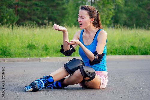Fototapeta Naklejka Na Ścianę i Meble -  attractive young athletic slim brunette woman in pink shorts and blue top with protection elbow pads and knee pads on roller skates sitting on the asphalt in the park . fall concept