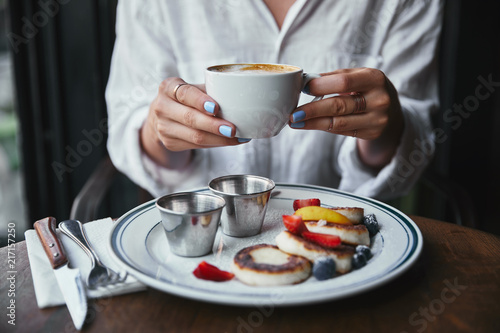 cropped shot of woman holding cup of coffee at restaurant above cheese pancakes on table
