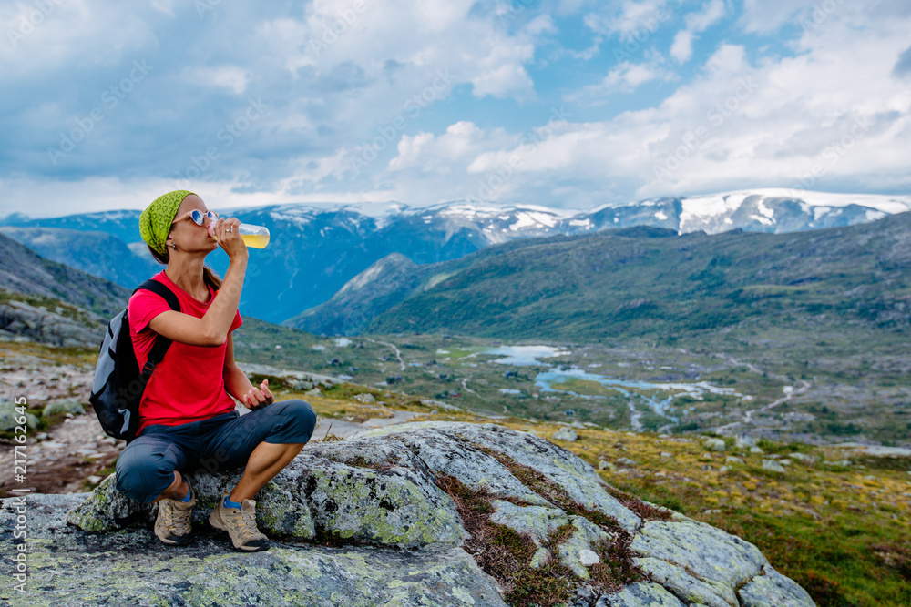 Naklejka premium Travel in Norway, woman hiker in bright sportswear with backpack sitting on big stone, relaxing and drinking water over scandinavian summer view.