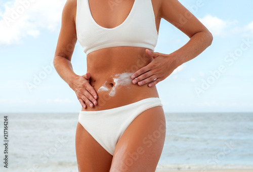 Beautiful woman on the beach in white swimsuit is applying sunscreen cream
