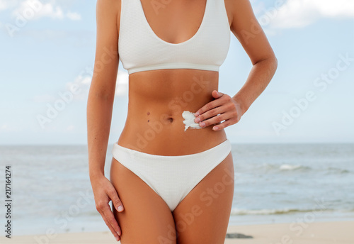 Beautiful woman on the beach in white swimsuit is applying sunscreen cream