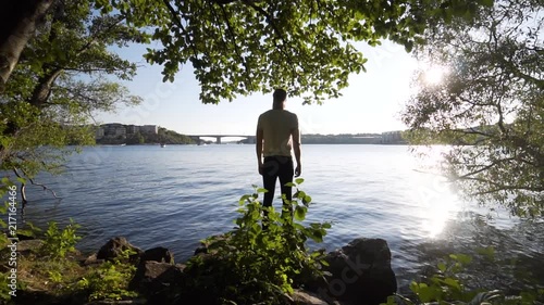 A tall Caucasian male stands by a canal in Stockholm Sweden. Steady glide cam footage moving towards the model in slow motion. Beautiful sunny summer day, green trees and a blue sky. photo