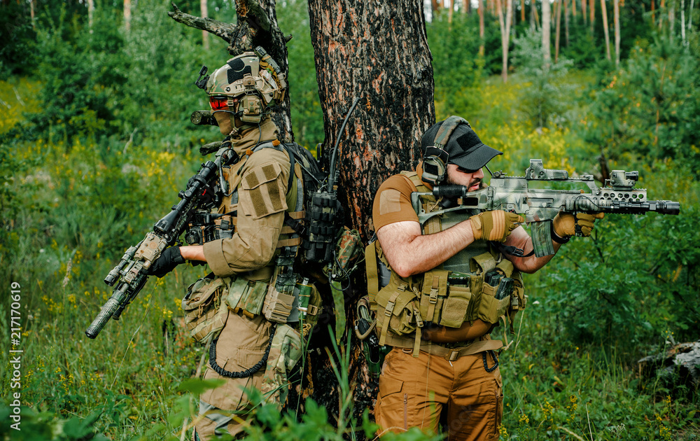 Airsoft men with guns stand back to back. Soldiers stand on hill and on call in the forest