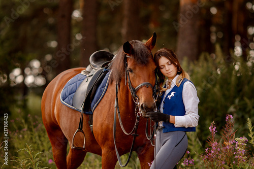 Young woman rider equestrian stands next to brown horse in forest.