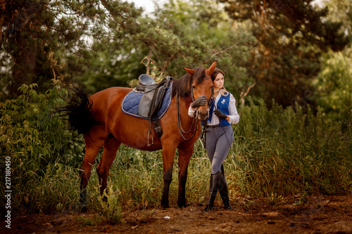 Young woman rider equestrian stands next to brown horse in forest.