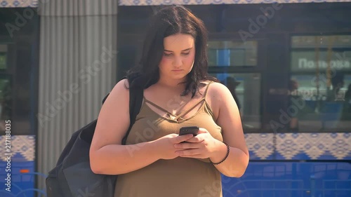 Young serious beautiful obese woman typing on phone and standing on station, train in background going away photo