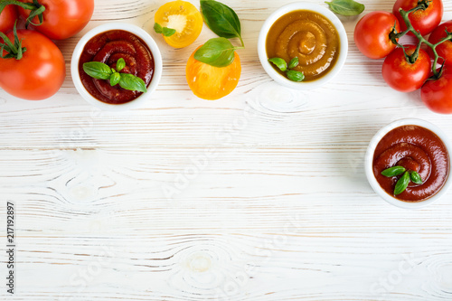 Tomato ketchup sauce in a bowls with basil and tomatoes on white wooden table. Different kinds of ketchup. Top view. Copy space