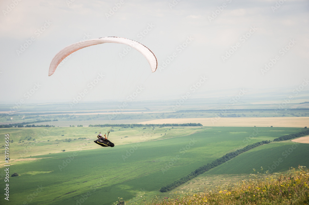 White orange paraglide with a paraglider in a cocoon against the background of fields of the sky and clouds. Paragliding Sports White orange paraglide with a paraglider in a cocoon against the background of fields of the sky and clouds. Paragliding Sports