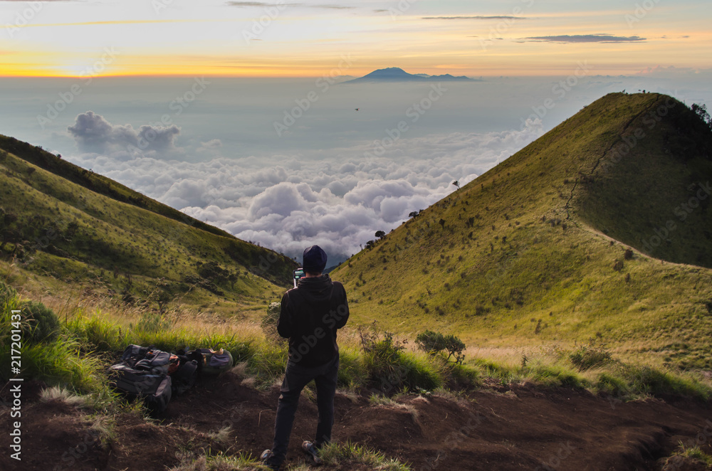 A drone pilot is flying quadrocopter on the mountain above the clouds. Sunrise at mount Merbabu, Indonesia, above the clouds. Volcano Lawu is at the background. Shooting aerial footage.