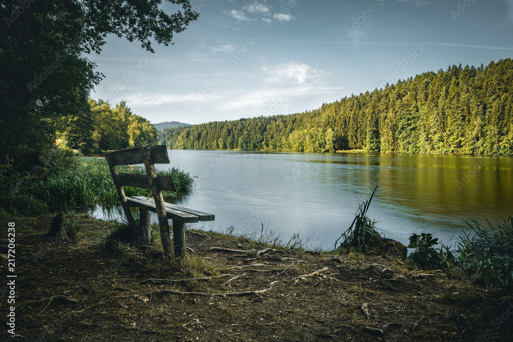 Bench in front of a lake with clouds on the sky and trees behind in the bavarian forest