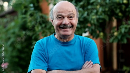 Charming handsome senior man in casual blue shirt keeping arms crossed and smiling while standing on formal garden. At first he is sad, but after hearing a joke begin to smile