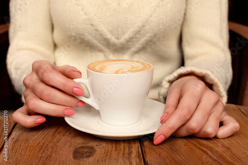 Lonely woman with nice manicure in warm wood sweater drinking coffee in the morning, view of female hands holding cup of hot beverage on wooden desk, retro toned.