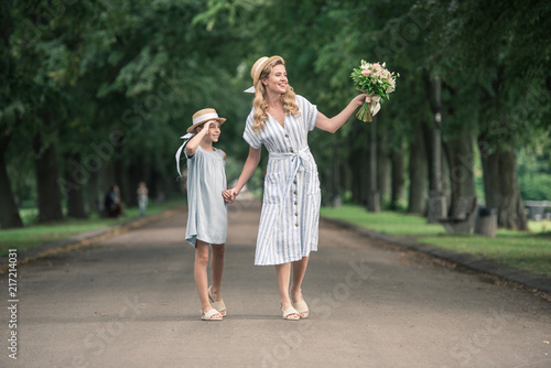 mother and daughter in straw hats with bouquet holding hands and walking on path in green park