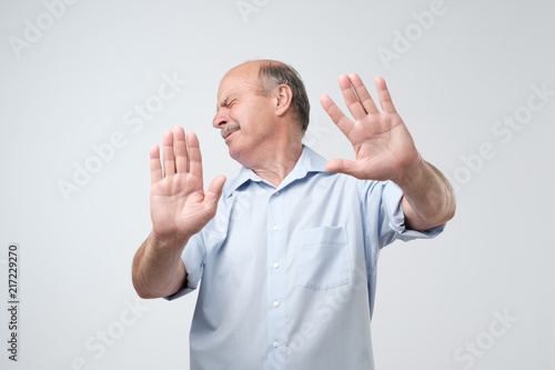 Displeased mature man refusing, stretching hands to camera over grey background.