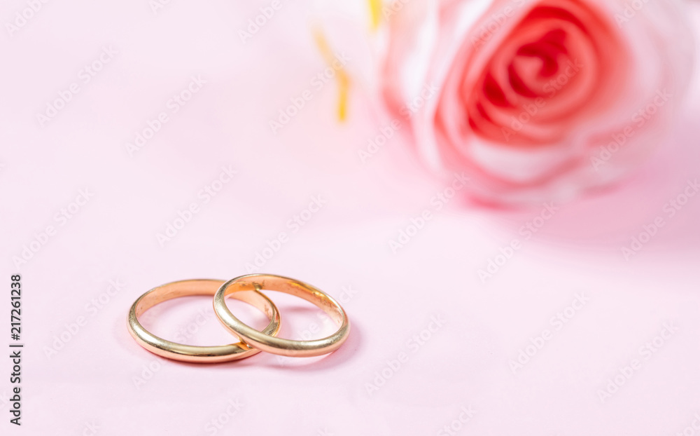 Close up view of golden wedding rings and a blurry pink rose, copy space, on a pink background. Close up view of golden wedding rings and a blurry pink rose, copy space, on a pink background.
