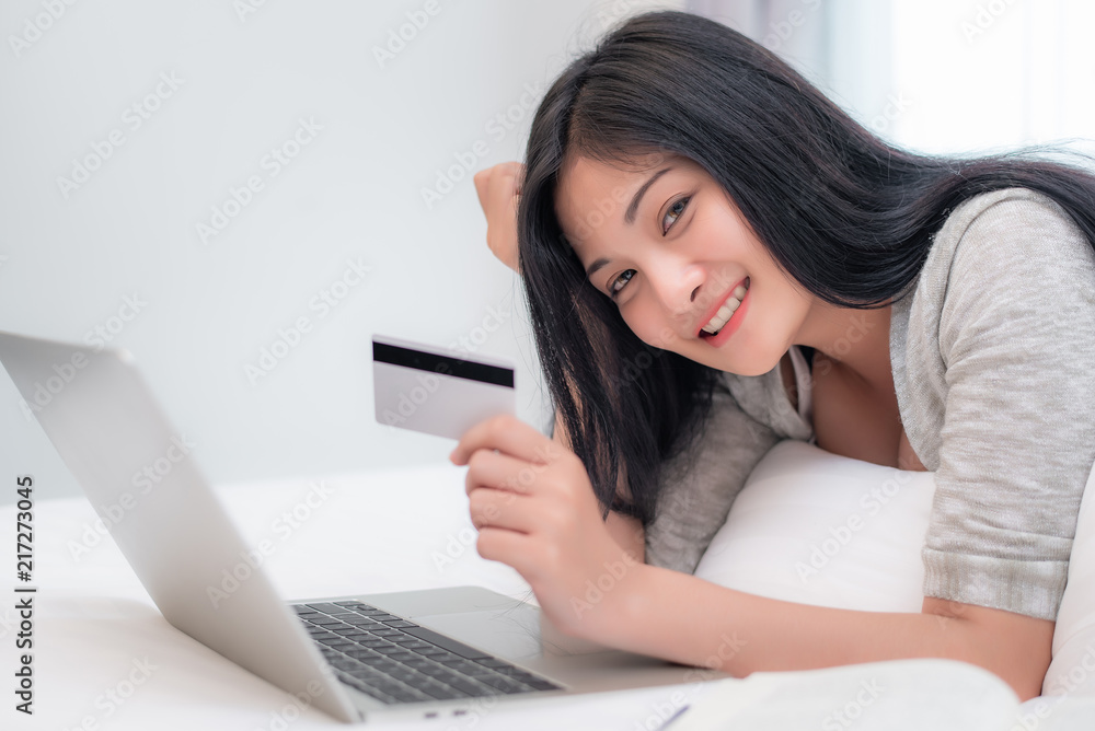 Pretty attractive Asian woman shopping online by laptop with credit card, laying on the bed at bedroom Pretty attractive Asian woman shopping online by laptop with credit card, laying on the bed at bedroom