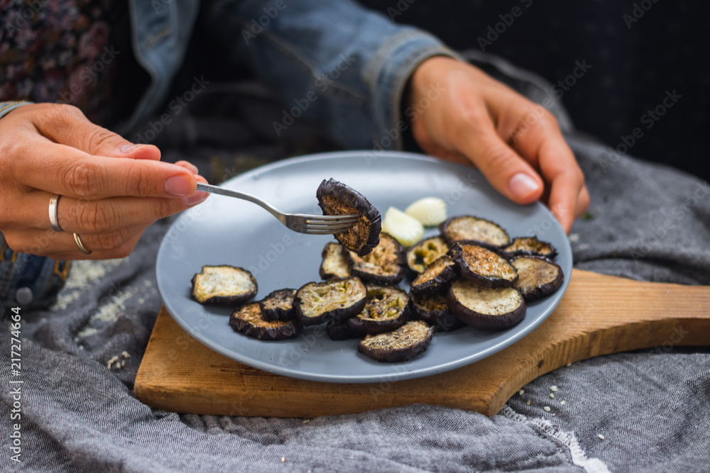 Woman hands sticks on fork roasted eggplants slices fro stylish grey plate. Sprinkled with herbs, black pepper and garlic powder. Perfect vegan dish. No refined oil. Vegetarian lunch. Small portion Woman hands sticks on fork roasted eggplants slices fro stylish grey plate. Sprinkled with herbs, black pepper and garlic powder. Perfect vegan dish. No refined oil. Vegetarian lunch. Small portion