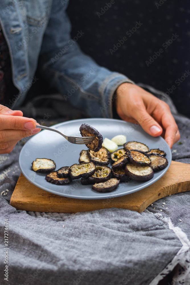 Woman hands sticks on fork roasted eggplants slices fro stylish grey plate. Sprinkled with herbs, black pepper and garlic powder. Perfect vegan dish. No refined oil. Vegetarian lunch. Small portion