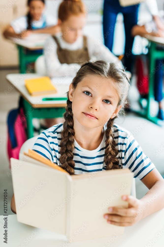 beautiful little schoolgirl with book looking at camera during lesson at school beautiful little schoolgirl with book looking at camera during lesson at school