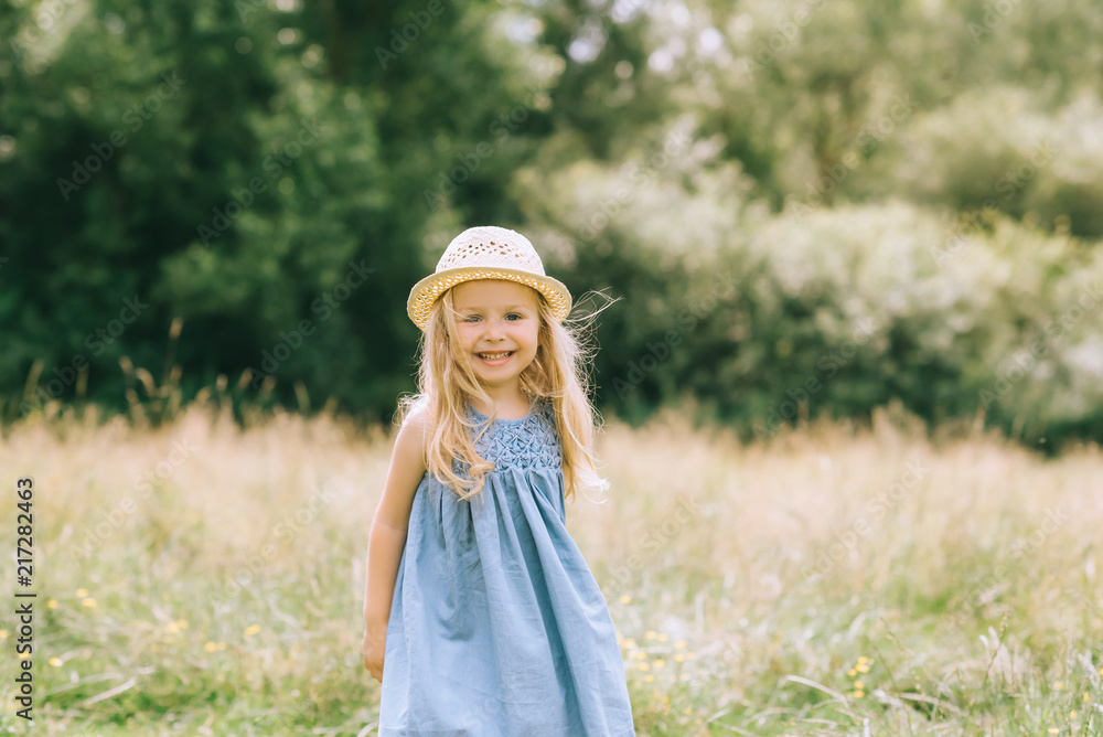 adorable blonde smiling child in dress and straw hat in field