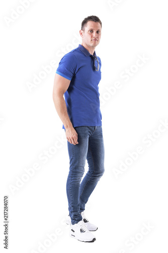 Side view of relaxed young adult casual man walking with sunglasses on neck looking at camera. Full body isolated on white background. 