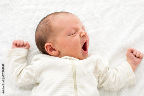 From above of newborn baby in white overall sleeping and yawning adorably lying on white blanket