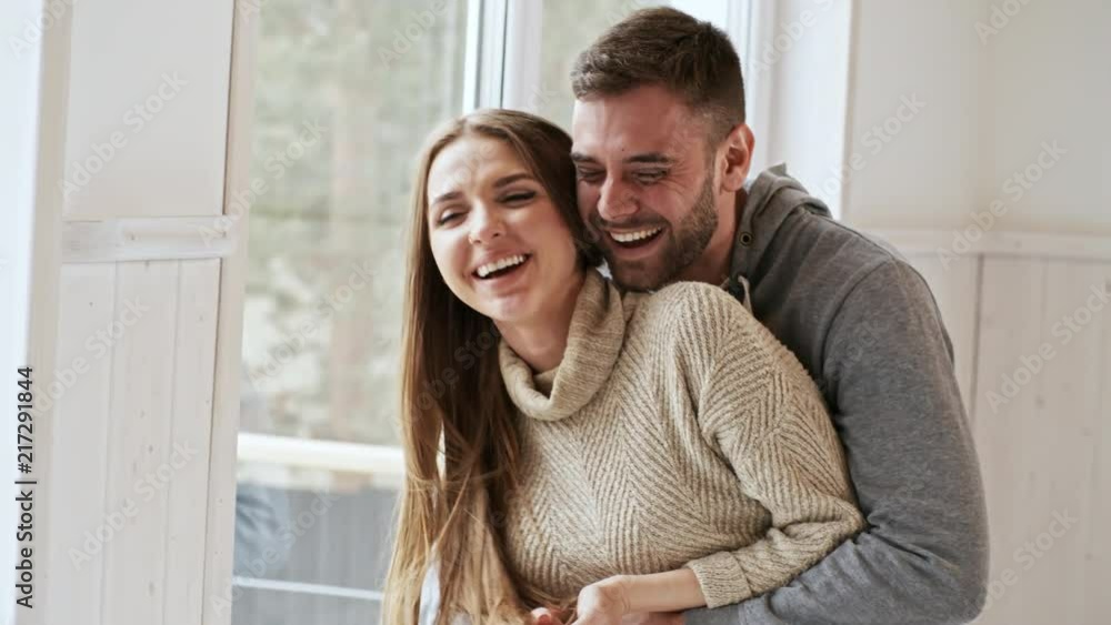 Handheld shot of happy young couple laughing and hugging by the window Handheld shot of happy young couple laughing and hugging by the window