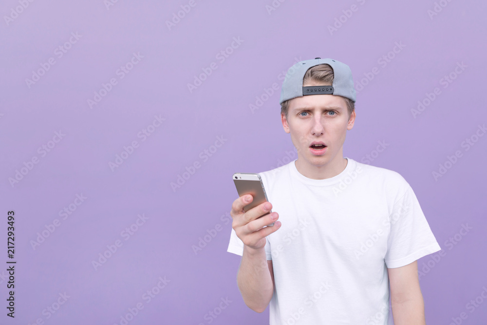 Amazed young man in a white T-shirt stands with a smartphone in his hands on a purple background and looks at the camera. Portrait of a student using a smartphone against a purple background. Amazed young man in a white T-shirt stands with a smartphone in his hands on a purple background and looks at the camera. Portrait of a student using a smartphone against a purple background.