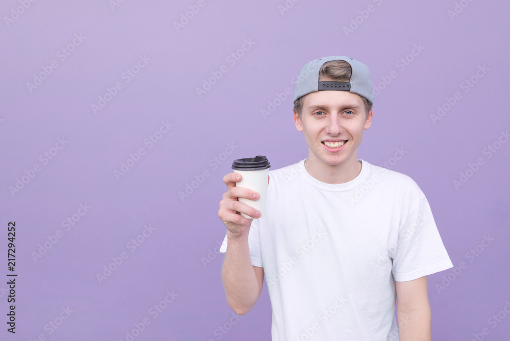 Portrait smiling young pretty man in a white t-shirt standing with a glass of coffee on a purple background and looking at the camera.Happy student with a coffee on the background of a purple wall Portrait smiling young pretty man in a white t-shirt standing with a glass of coffee on a purple background and looking at the camera.Happy student with a coffee on the background of a purple wall