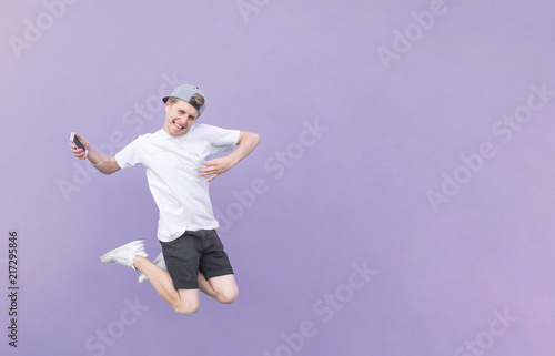 Portrait of a young man jumping with headphones and a smartphone on a pastel purple background. The teenager jumps with headphones in his ears and a smartphone in his hand