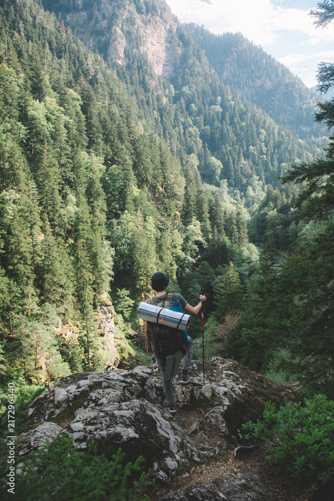  a young guy with a tourist backpack looks down from the mountain to the river and a thick green forest