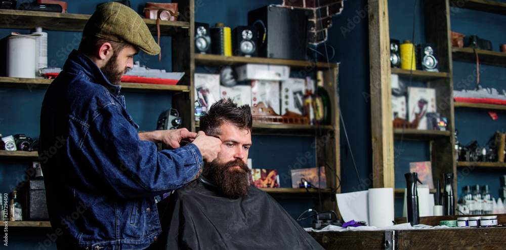 Hipster client getting haircut. Barber with hair clipper work on hairstyle for hipster, barbershop background. Hipster lifestyle concept. Barber with clipper trimming hair on temple of bearded client