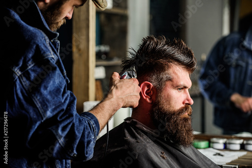 Barber with hair clipper works on hairstyle for bearded man barbershop background. Barber styling hair of brutal bearded client with clipper. Hipster lifestyle concept. Hipster client getting haircut