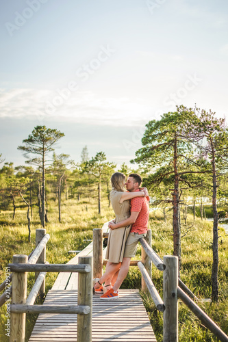 side view of affectionate couple hugging on wooden bridge with green plants and blue sky on background