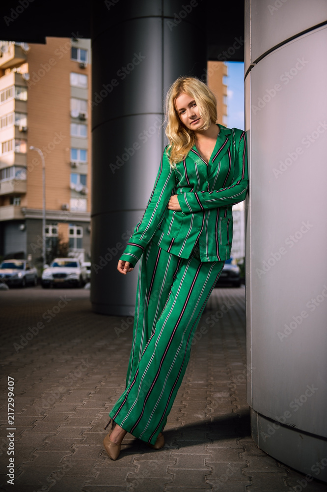 Beautiful girl in a green costume standing near a gray column. Stylish blonde. Fashionable girl. Everyday life of a beautiful girl. Lovely blonde in a stylish suit. Gray wall.
