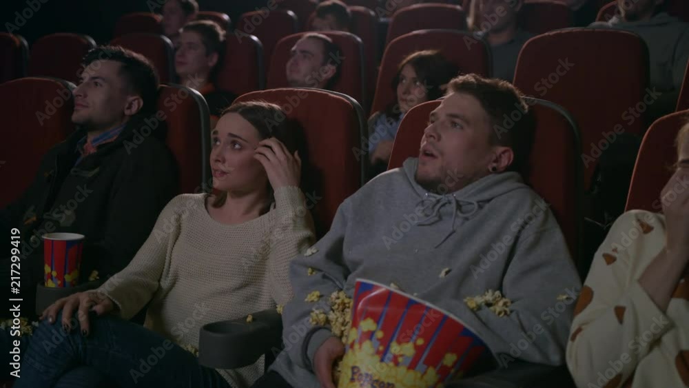 Young guy scared while watching horror film in movie theatre. Young man sprinkling popcorn in slow motion at cinema. Spectators frightened by horror film. People watching scary movie Young guy scared while watching horror film in movie theatre. Young man sprinkling popcorn in slow motion at cinema. Spectators frightened by horror film. People watching scary movie