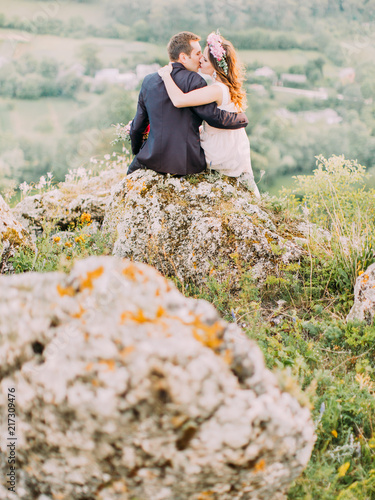 Back view of the kissing newlyweds while sitting on the rock. photo