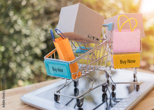 Mini Cardboard box, shopping bag in trolley with word buy now on tablet. Consumers can buy products directly from seller over internet using web browser. Ideas about online shopping and e-commerce.