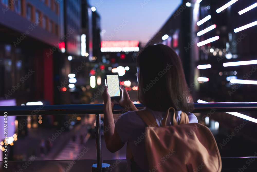 rear view of woman with backpack and smartphone with map on screen in hands standing on night city street rear view of woman with backpack and smartphone with map on screen in hands standing on night city street