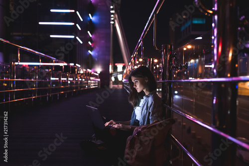 side view of young woman using laptop on street with night city lights on background
