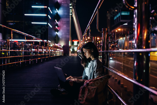 side view of young woman using laptop on street with night city lights on background