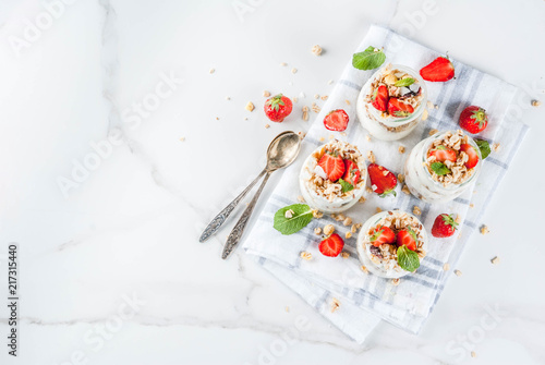 Healthy summer breakfast idea, homemade layered parfe dessert in small jar with granola, yoghurt and strawberry, dark background copy space above photo