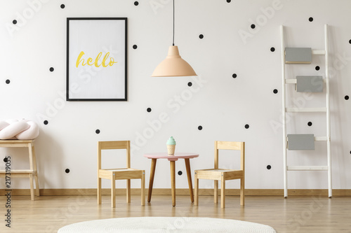 Pastel pink pendant above wooden play dining set for kids and books on a ladder in a cute preschool room interior with black and white polka dot wallpaper
