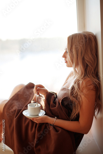 Young beautiful Girl drinking coffee sitting near the window in morning. Blond sexy naked female Boudoir photo standing in front of large window wrapped in blanket with her back to camera