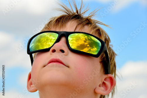 A little boy looks at the cloudy sky with glasses. The clouds are reflected in the glass. Yellow glasses against the blue sky.