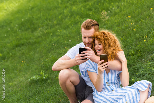 happy redhead couple with smartphones sitting on grassy meadow