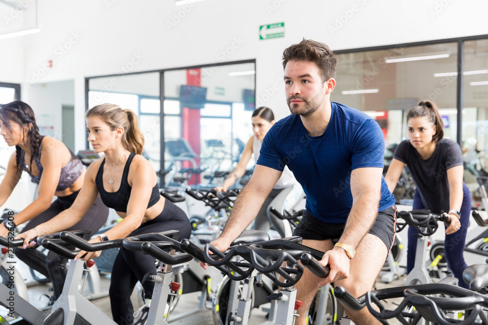 Man Riding Stationary Bicycle With Friends In Gym Man Riding Stationary Bicycle With Friends In Gym