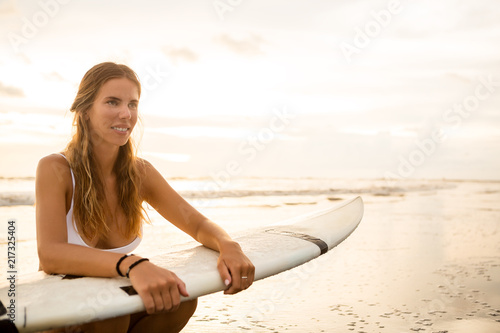 Young surfer girl in sexy white bikini with blank surfing shortboard sit in water at sunset ocean. Modern family lifestyle, people water sport adventure camp and extreme swim on summer vacation. © Mikalai Bachkou